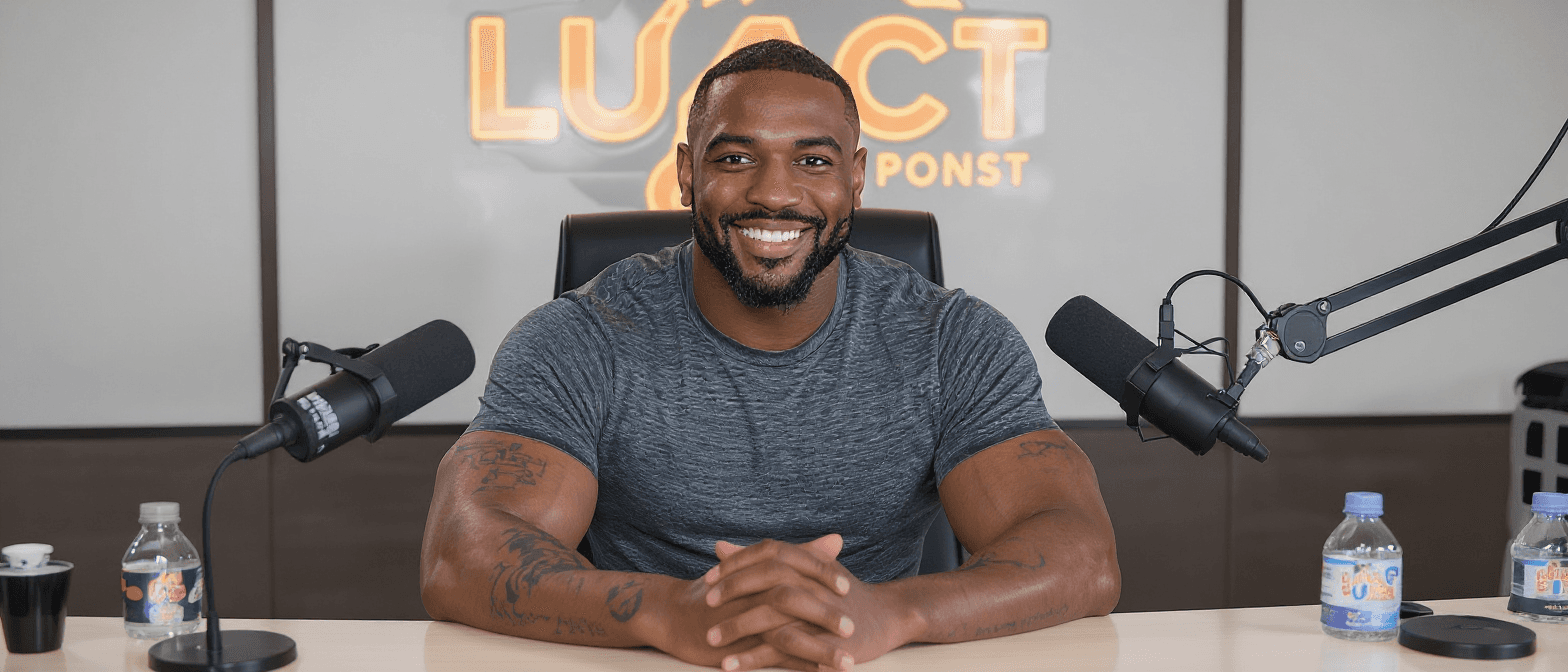 A high-resolution 1568x672 image of a smiling Black man sitting at a podcast desk with his hands clasped. He wears a grey t-shirt. Two microphones flank him, and water bottles sit on the desk. This image was generated using the Flux Kontext Pro model.